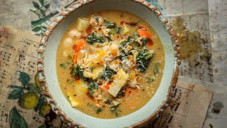 A rustic ceramic bowl filled with kale white bean soup featuring carrots, kale, potatoes, beans, and shredded cheese on top, set on a surface decorated with sheet music and colorful illustrations.